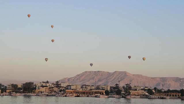 Landscape photo. We're looking across the pale waters of the Nile at the thin band of a town with a wild mix of light coloured houses with dark trees growing in between. Behind it grows a mountain range, looking bare and red in the backlight of the sun rising to the left outside of the photo. There's the wide expanse of the pale blue morning sky with a handful of scattered hot air balloons rising silently into the sky to watch the sunrise.