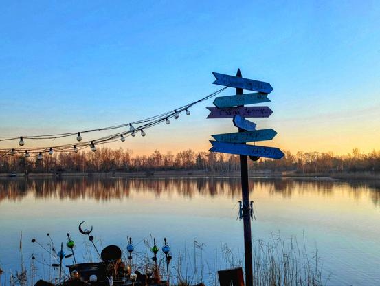 A scenic view of a calm lake at sunrise, with a wooden signpost in the foreground pointing in multiple directions. The sky transitions from a deep blue at the top to a warmer orange and yellow near the horizon. Bare trees line the far shore of the lake, their reflections visible in the still water. To the left, a string of vintage-style light bulbs hangs diagonally across the frame. In the lower left corner, there's a glimpse of decorative garden ornaments. The overall atmosphere is tranquil and picturesque.