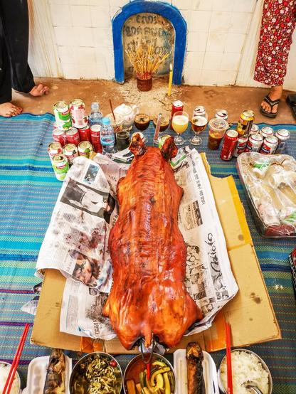 Photo shows a set up of food and drinks offering in front of a horizontal grave. The main item is a full-size BBQ-pork. It is surrounded by offerings of fruit, cake, juice, water, beer, and utensils. The offerings are placed on a picnic mat. The grave has a blue door and a front in a half-moon. The front is white, and the outline is red.