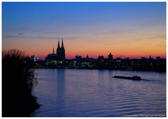 Skyline von Köln im Sonnenuntergang. Man erkennt den Kölner Dom und die angrenzenden Kirchen und Häuser im Halbdunkel entlang des Rheins. Der Himmel ist von tiefen Blau bis zum Horizont in zarten Rot. Die Gebäude sind von scharfem Kontrast, im Vordergrund stehen Büsche am Ufer und ein Schiff zieht in Richtung Dom über den Rhein.