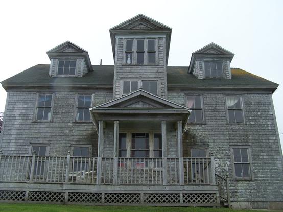 A very old run-down building, resembling a hotel in a horror movie. It has a set of pillars holding a roof over the entrance, and above, another tower rises up in front of the whole building  Each roof of the wide building has two dormer windows. 
And long windows on each side of the front entrance tower
A wooden deck with rails, runs from left to past the entrance