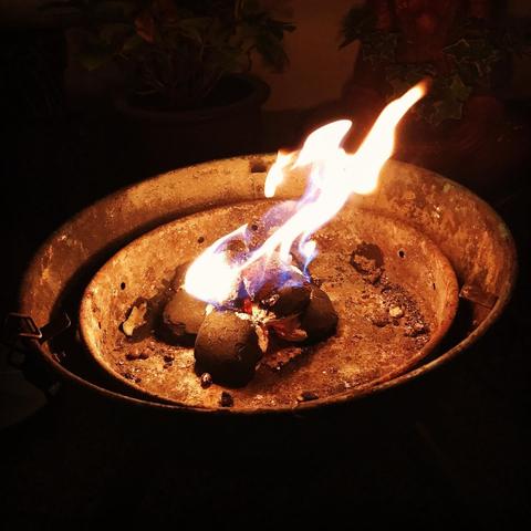 Das Bild zeigt eine Flamme, die in einem runden, rostigen Metallbehälter brennt. Die Form der Flamme erinnert an einen springenden Hasen. Der Behälter scheint aus Metall zu sein und hat eine runde Form mit einem offenen Rand. Im Inneren des Behälters sind mehrere schwarze Kohlen sichtbar, die in Flammen stehen. Die gesamte Szene ist in einem dunklen Umfeld aufgenommen, wodurch die Flamme besonders hervorsticht.