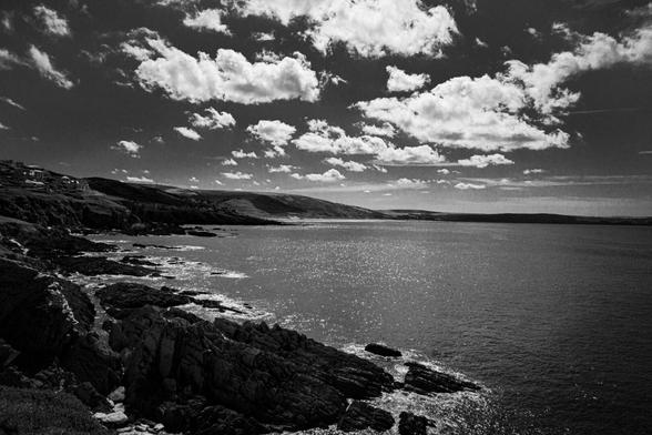 Cornwall landscape. Sunlit clouds drift across the sky, their reflections shimmering on the ocean’s surface. The rocky coastline and rolling hills create a dramatic black-and-white scene, emphasizing the rugged beauty of nature.