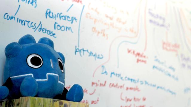 A godot plushie, sitting in front of a whiteboard wall with an idea mind-map scribbled on