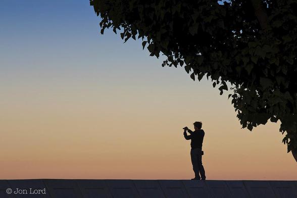 A Simple Colour Dawn Photo In Landscape Format. At The Base Of The Image Stretching Across From Left To Right And Rising Only About Ten Per Cent Is A Concrete Sea Wall. Standing On The Sea Wall, A Little To The Right Of Centre Is A Man, Camera In Hand, Taking A Photo Of The Sunrise Scene. Above The Man And Running Diagonally From The Lower Right Corner Towards The Upper Left Corner Are The Branches And Leaves Of A Large Tree, Nearly All In Shadow With Only The Green Showing In The Foliage In A Few Places. In The Background Is A Clear And Cloudless Sky, The Colours Graduating From A Rich Orange At The Bottom Through Yellow To Blue At The Very Top Of The Image. 
The Location: Redondo Beach, California (2011).