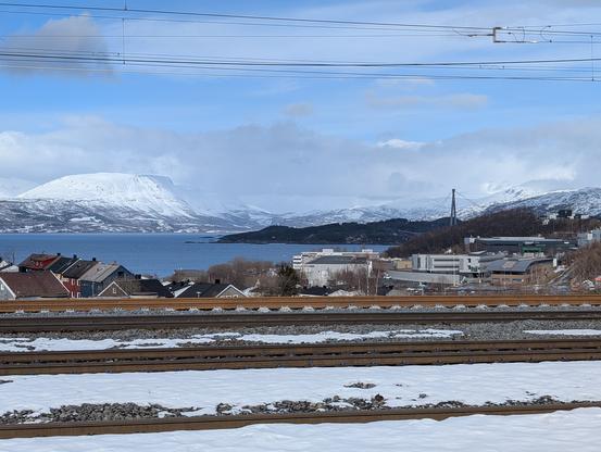 View from narvik station. Snow covered tracks in the front, snow covered mountain in the back left and in the far right the bridge. Some houses in between.