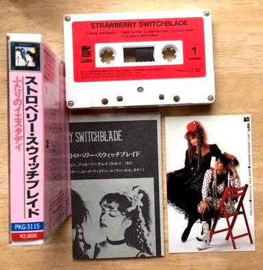 The contents of the Strawberry Switchblade album's Japanese cassette release l;aid out on a table. There is the spine of the cassette insert card, a folded monochrome info sheet, a cassette, and a small colour photo of the duo.