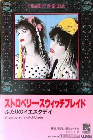 The cover of the Strawberry Switchblade album's Japanese cassette release. The album artwork is a colourised photo of Jill and Rose in polka dot dresses with plastic flowers in their hair from which ribbons are trailing. It is bordered with a stylised floral pattern. Below the artwork is a pink area with Japanses lettering.