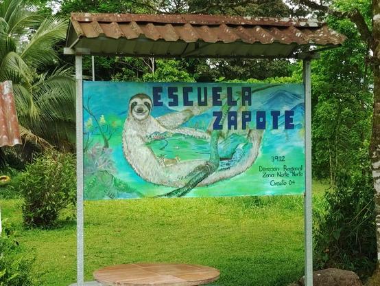 Eine Art Gemälde, das ein Faultier zeigt, das eine bergige Landschaft umarmt. Es steht "Escuela Zapote" darauf. Es steht auf zwei dünnen Metallpfeilern, darüber ein kleines Dach zum Schutz vor Regen.
Im Hintergrund viel Grün und eine Kokospalme.