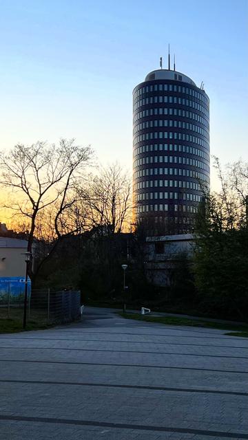 Ein modernes, rundes Hochhaus mit dunkler Glasfassade ragt in den Himmel bei Sonnenuntergang. Die untergehende Sonne taucht den Himmel in warme Orange- und Blautöne. Im Vordergrund führt ein gepflasterter Weg zwischen Bäumen und einem Zaun entlang. Die kahlen Äste der Bäume bilden ein filigranes Muster vor dem leuchtenden Abendhimmel. Eine Laterne steht am Wegrand und verstärkt die urbane Atmosphäre.