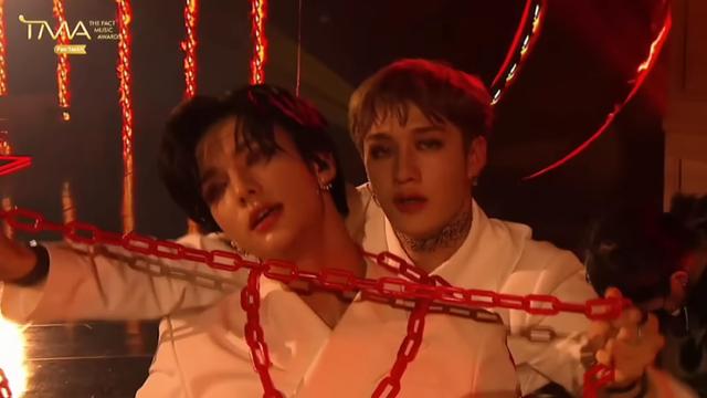 Hyunjin and Bang Chan performing Red Lights t an awards show. Hyunjin's leaning back against Chan, who's holding a red chain up in front of him.