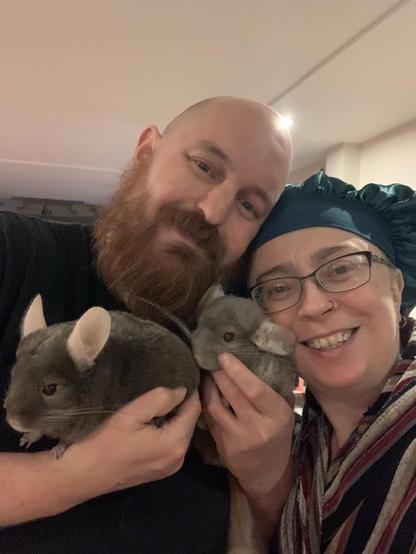A smiling couple poses for a selfie, holding two gray chinchillas. The man has a beard and is wearing a black shirt, while the woman, wearing glasses and a turquoise headscarf, is smiling brightly.