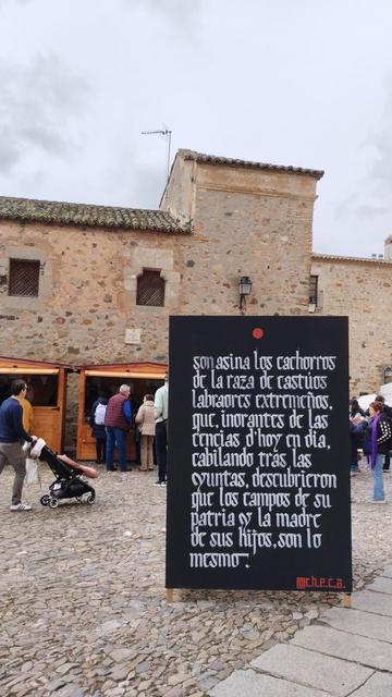 Una calle empedrada con edificios antiguos (es el casco antiguo de Cáceres) y hay puestos de artesanía en cabañas de madera. En medio de la foto con fondo negro y letras blancas (y como góticas) está escrito un fragmento de El miajón de los castúos de Luis Chamizo. Es obra de une artista que se llama @c.h.e.c.a.