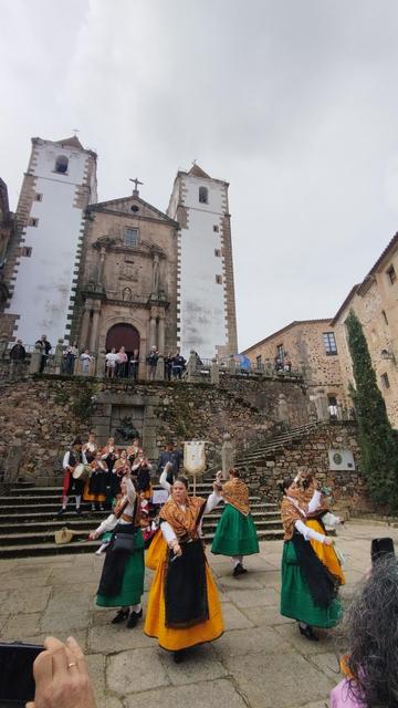 En la parte superior de la imagen se ve una iglesia con dos torres encaladas a cada lado, bajando una escalinata de piedra doble y un tramo de escalera con gente tocando y cantando mientras en el primer plano bailan jotas. Llevan indumentaria tradicional extremeña