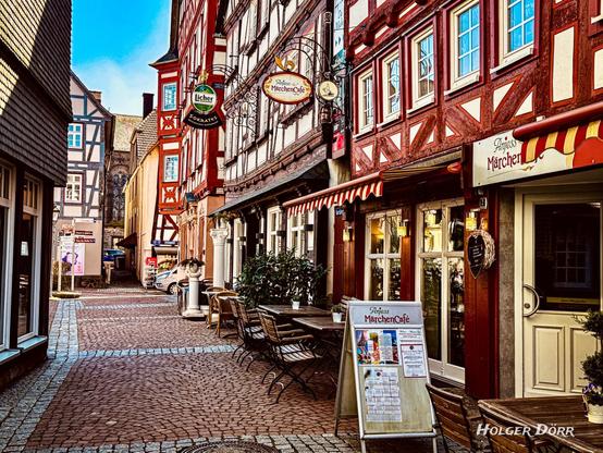 Romantische Fachwerkgasse in der Altstadt von Grünberg mit dem liebevoll dekorierten „Anjess MärchenCafé“. Vor dem Café stehen einladende Tische auf dem Kopfsteinpflaster, überragt von kunstvoll verzierten Fassaden und nostalgischen Wirtshausschildern. Im Hintergrund ragt eine alte Kirche empor – eine Szenerie wie aus einem Märchenbuch.