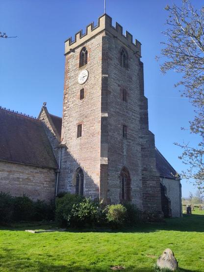 a lush green meadow in which stands a mediaeval church with a Victorian rebuilt tower. it is in sunshine and a bright blue cloudless sky.