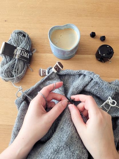 ENG Shown from above are two hands holding circular knitting needles. The person is knitting sleeves on a grey cardigan. The project is resting on a table next to a blue heart-shaped mug with black coffee, two black needle stopper and a row counter

DEU Das Bild ist aus der Ich-Perspektive aufgenommen und zeigt die Hände einer Person, die mit Rundstricknadeln die Ärmel eines grauen Cardigans strickt. Das Strickprojekt liegt auf einem Holztisch und daneben sind eine blaue herzförmige Tasse mit schwarzem Kaffee, zwei Nadelstopper und ein Reihenzähler zu sehen.