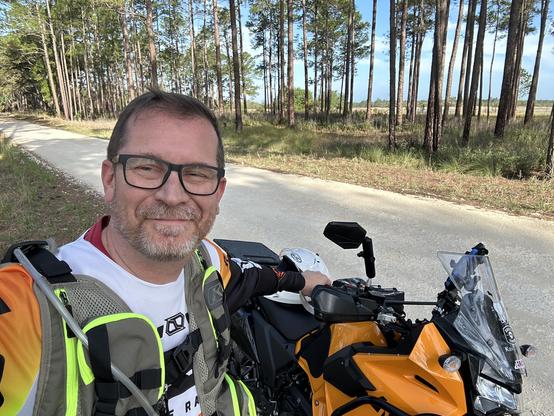 Me and my motorcycle, an orange KLR 650. Parked just off a dirt road, with a background of trees. The prairie can be seen through the gaps in the trees.