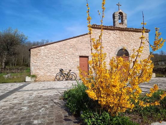 Chiesetta di campagna con la facciata realizzata con grosse pietre, con un piccolo campanile sormontato da una croce. Alla chiesa è poggiata una bicicletta, in primo piano una pianta ricca di fiori gialli nei colori della primavera.