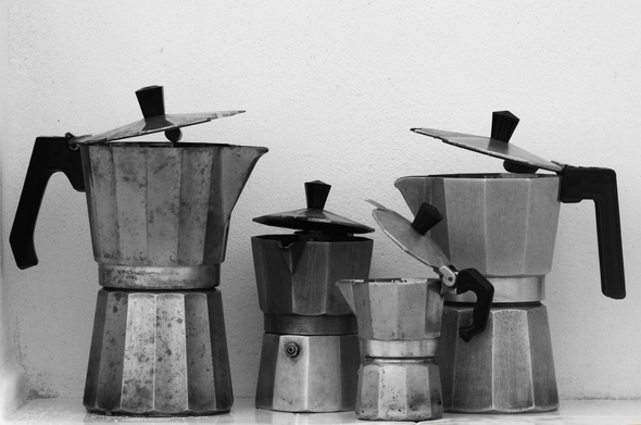 Fotografía en blanco y negro donde se ven cuatro cafeteras italianas antiguas y de distintos tamaños, con sus tapas entreabiertas, colocadas de manera que parece que están hablando entre ellas. El fondo es una pared blanca con gotelé, por lo que se aprecia la textura rugosa.