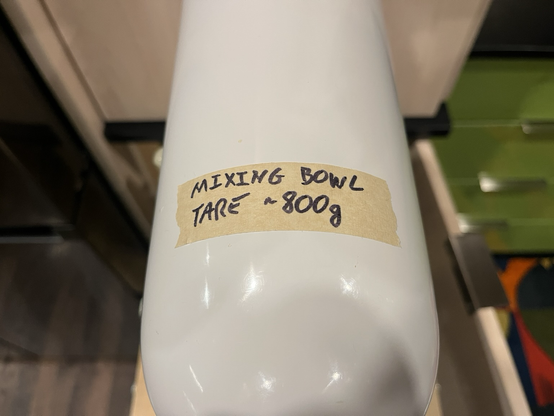The top of a white stand mixer with masking tape on it. In all caps, in handwritten black sharpie it reads “MIXING BOWL TARE ~800 g”.
