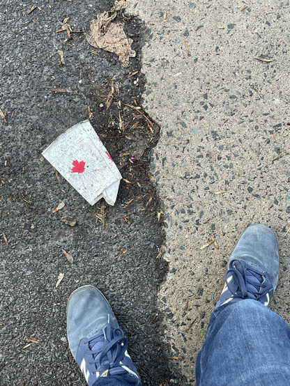 On a sidewalk, the photographer’s feet are in the frame with a used white cardboard cup from a fastfood  chain showing a canadian red maple leaf logo.