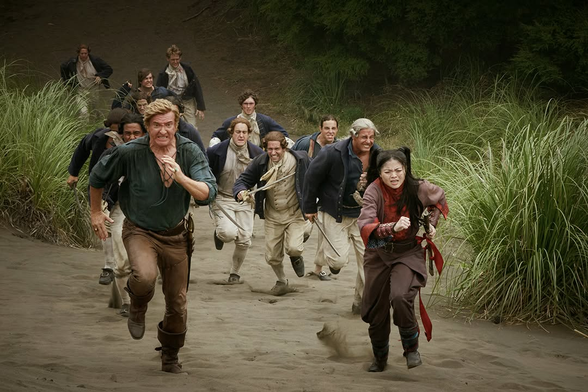 A still from the Our Flag Means Death Season 2 finale: Stede (Rhys Darby) and Zheng (Ruibo Qian) run from a group of English navy officers on a beach.