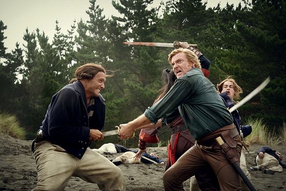 A still of Stede and Zheng fighting the English: Stede has gut-stabbed one of the officers, who is staring at him with a creepy grin, while Stede himself is grimacing and looking at something out of frame as he tries to pull his sword free. Behind him, Zheng is fighting another officer, her bloodied sword raised above her head.