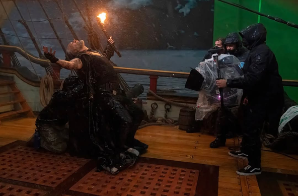 A behind-the-scenes photo from the storm scene in season 2, episode 2: Taika Waititi (Blackbeard) stands in the pouring “rain” on the deck of the Revenge set, bending backwards with his head tilted back and his arms stretched out, holding a lit torch in one hand. He is leaning on a barrel or similar prop, behind which a person in a black raincoat kneels on the ground to support Taika's back with both hands. Facing him are several camera crew and the camera itself, all wrapped up in protective rain gear. The huge LED screen used as a backdrop for the scene shows a stormy sea at night.