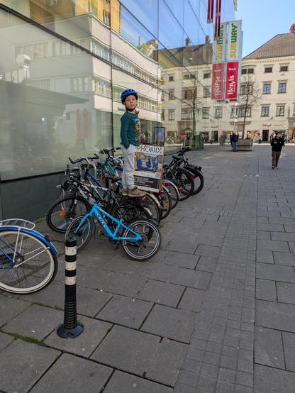 Ein Kind balanciert auf dem Gepäckträger eines Fahrrads, das bei einem Fahrradständer steht.