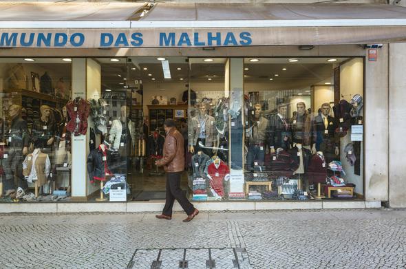 A man passes in front of a clothing shop window, his head is turned away from the camera and looking inside the shop. The window features male mannequins on the right side, the person in the middle in front of the entrance, and female mannequins on the left. The shop name is above the entrance, Mundo das Malhas.
