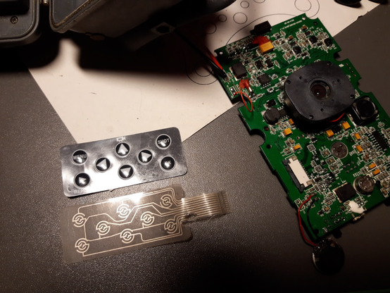 Auf einem Tisch liegt die blanke Platine einer Wildkamera. Daneben eine folienartige Button-Matrix und die darunter liegende flexible Leiterplatte. Bei der flexiblen Leiterplatte ist ein Loch in den Kunststoff gebrannt.