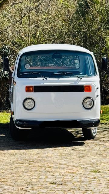 Das Bild zeigt eine klassische weiße Volkswagen-Bus-Van aus der Mitte des 20. Jahrhunderts. Die Van steht auf einem gepflasterten Weg vor einem Hintergrund aus Bäumen und Grün. Die Fahrzeugfront ist klar zu sehen, mit den charakteristischen runden Scheinwerfern und den orangefarbenen Blinkern. Der Kühlergrill ist schwarz und das VW-Logo ist deutlich sichtbar. Die Windschutzscheibe ist groß und rechteckig, mit zwei Wippern. Die Van hat einen klassischen, runden Designstil, der für die 1960er und 1970er Jahre typisch ist. Die gesamte Fahrzeugoberfläche ist sauber und gut gepflegt, was auf eine sorgfältige Restaurierung oder Erhaltung hinweist.