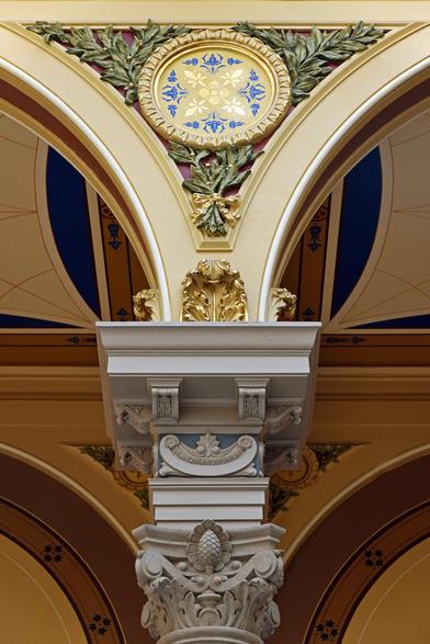 Color photo of the elaborate capital of an interior column from which two arches spring. A medallion occupies the space between the arches. Nearly all of the surfaces are embellished with designs painted in yellow, gold, blue, and green.