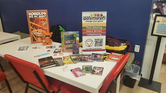 An assortment of 9th Level Games RPGs spread out on a table including Mazes, Savage Sisters, good dogs, sentai & Sensibility, scurvy buggers,  piegons 11, kobolds ate my baby, the excellents, Nancy Druid, and horseshoe academy. Two table signs in the background. One is of Kobolds with Creature the Kobold plushie proped on it. The other is for Polymorph.