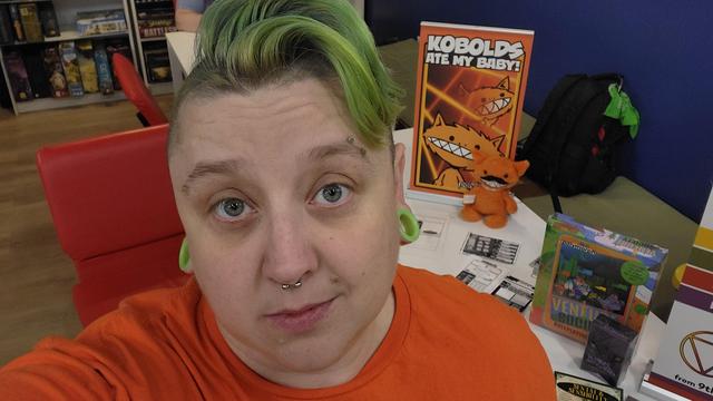 A green haired nerd in front of an orange kobolds ate my baby sign. Hanging out with Creature, the Kobold Plush.