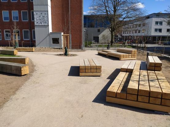 Das Bild zeigt Sitzmöbel aus Massivholz die in Gruppen an einem angelegten Parkweg aufgestellt sind. Zwei der Sitzgelegenheiten bieten große Plattformen auf denen man sich auch hinlegen kann. Daneben sind einige neugepflanzte Bäume und Anpflanzungen von Stauden zu erkennen.