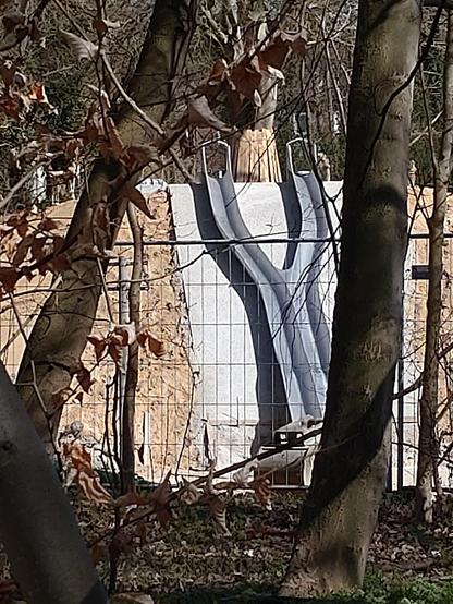 Das Bild zeigt eine lange Zwillingsrutsche zwischen zwei Bäume hindurch hinter einem Bauzaun. Die Zwillingsrutsche bildet eine Y Form. Zwei Einstiege etwa 2 m voneinander entfernt. Zum Auslauf hin werden beite Rutschbahnen zusammen geführt und verlaufen direkt nebeneinander.