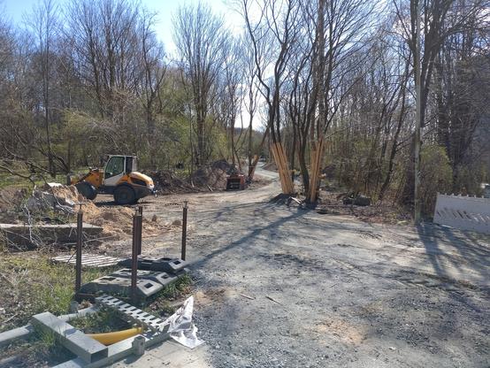 Blick auf eine Baustelle für einen Weg zwischen Bäumen hindurch. Der Weg ist durch ein Bett an Grobmaterial schon zu erkennen. Der Weg schlängelt sich in Kurven an Bäumen vorbei ins Gelände. Am Rand des Weges ist ein Radlader geparkt.