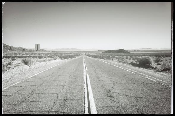 A long view of Route 66, shot from the center, in the glaring midday sun. The old, cracked asphalt highway runs off into the distance across low, scrubby desert marked only by a few baren hills on the left and right. 

There is no shade. There is no water. There are no people. There's no reason to be here.