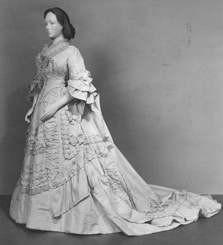 A black and white photo of a mannequin dressed in a light-colored gown with a pointed waistline, ruffled sleeves, and a train, the skirt heavily embellished with self-fabric trim.