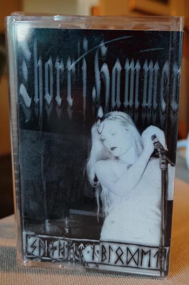 Cassette cover. Black and white photo of Runhild Gammelsæter on stage in white (in general, looking very pale/white because of the light), holding a microphone on stand, looking to (her) right). 

"Thorr's Hammer" above her.
Runes in the bottom, probably the name of the tape.