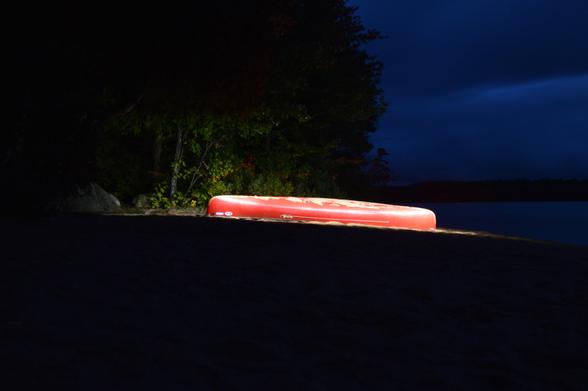 A brightly lit red canoe lies on it's side completely surrounded by darkness. Through the dimness of the dark you can see that it sits upon a beach at a lake's shore with trees behind. There is sand on the bottom of the canoe.