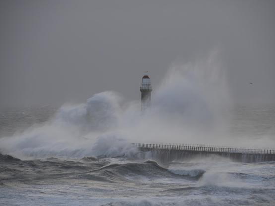 A photo of a lighthouse as a huge waves hits the pier on which it stands. The crashing wave obscures the lower parts of the lighthouse as the strong wind blows the spray.