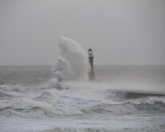 A photo of a lighthouse as a huge waves hits the pier on which it stands. The crashing wave obscures the lower parts of the lighthouse as the strong wind blows the spray.