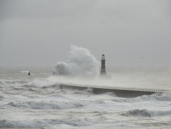 A photo of a lighthouse as a huge waves hits the pier on which it stands.