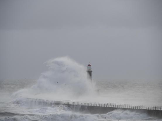A photo of a lighthouse as a huge waves hits the pier on which it stands. The crashing wave obscures the lower parts of the lighthouse as the strong wind blows the spray.