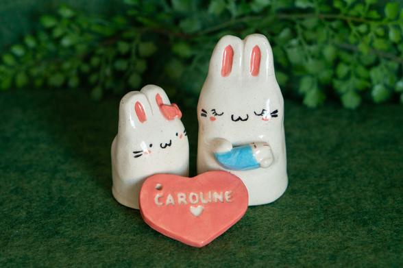 Two cute minimalistic white ceramic bunny figurines, about 3 inches tall, and a pink ceramic heart-shaped tag with the name Caroline stamped onto it. One figurine is a girl bunny with a pink bow on one ear, and the other is a slightly larger mama bunny holding a tiny baby bunny in a blue blanket.