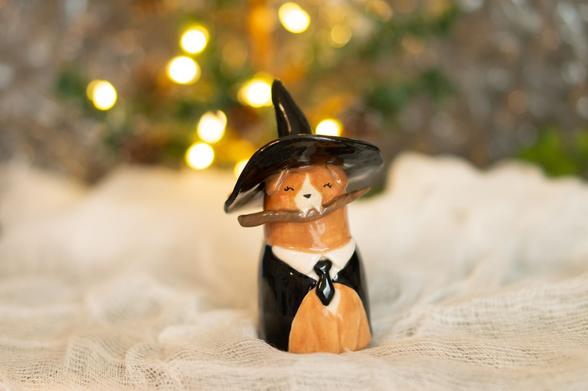 A cute ceramic puppy figurine, about three inches tall, wearing a witch's hat and black robes. The puppy is brown and has a mischievous smile while holding a brown magic wand between its teeth.
