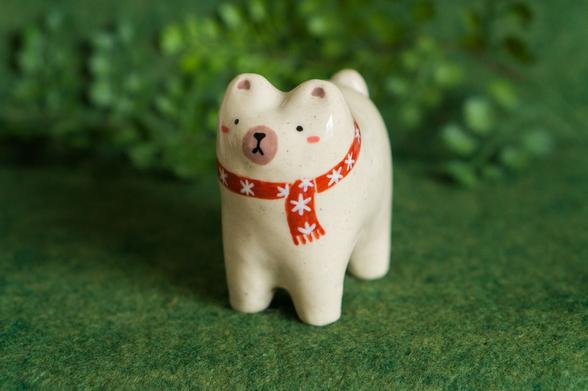 A cute minimalistic ceramic polar bear figurine in a standing position, wearing a red winter scarf around its neck. It has beady eyes and a simple downturned mouth like a classic teddy bear might have.